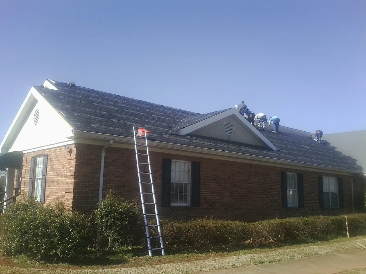 Crew tearing shingles off a brick ranch roof with a ladder set — Emergency Roof Repair work by our crew in Bonduel