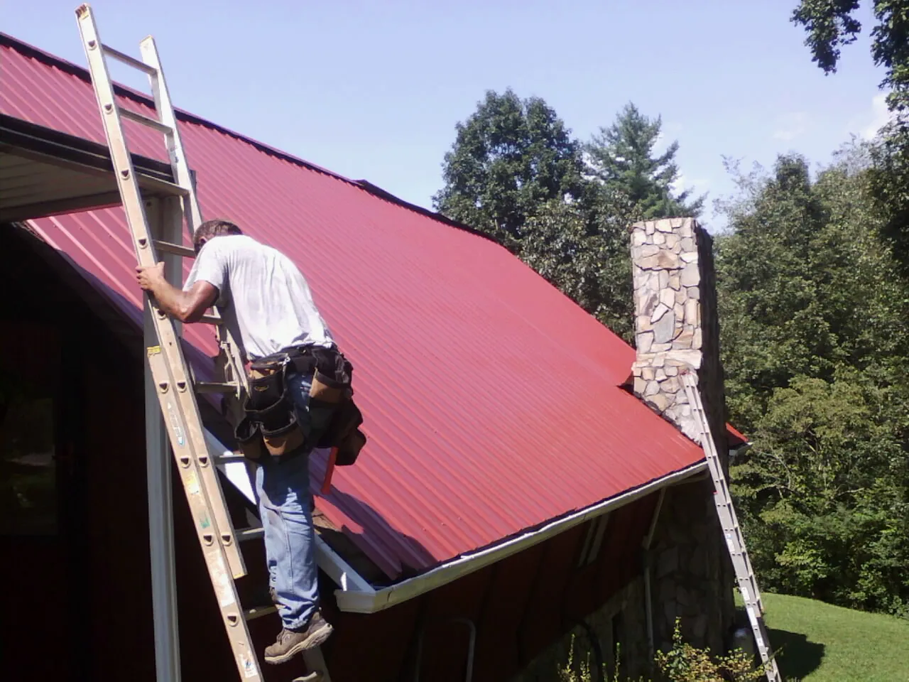 Roofer on a ladder beside a red metal roof and stone chimney — Soffit & Fascia Repair by Roofing Brothers in Bonduel